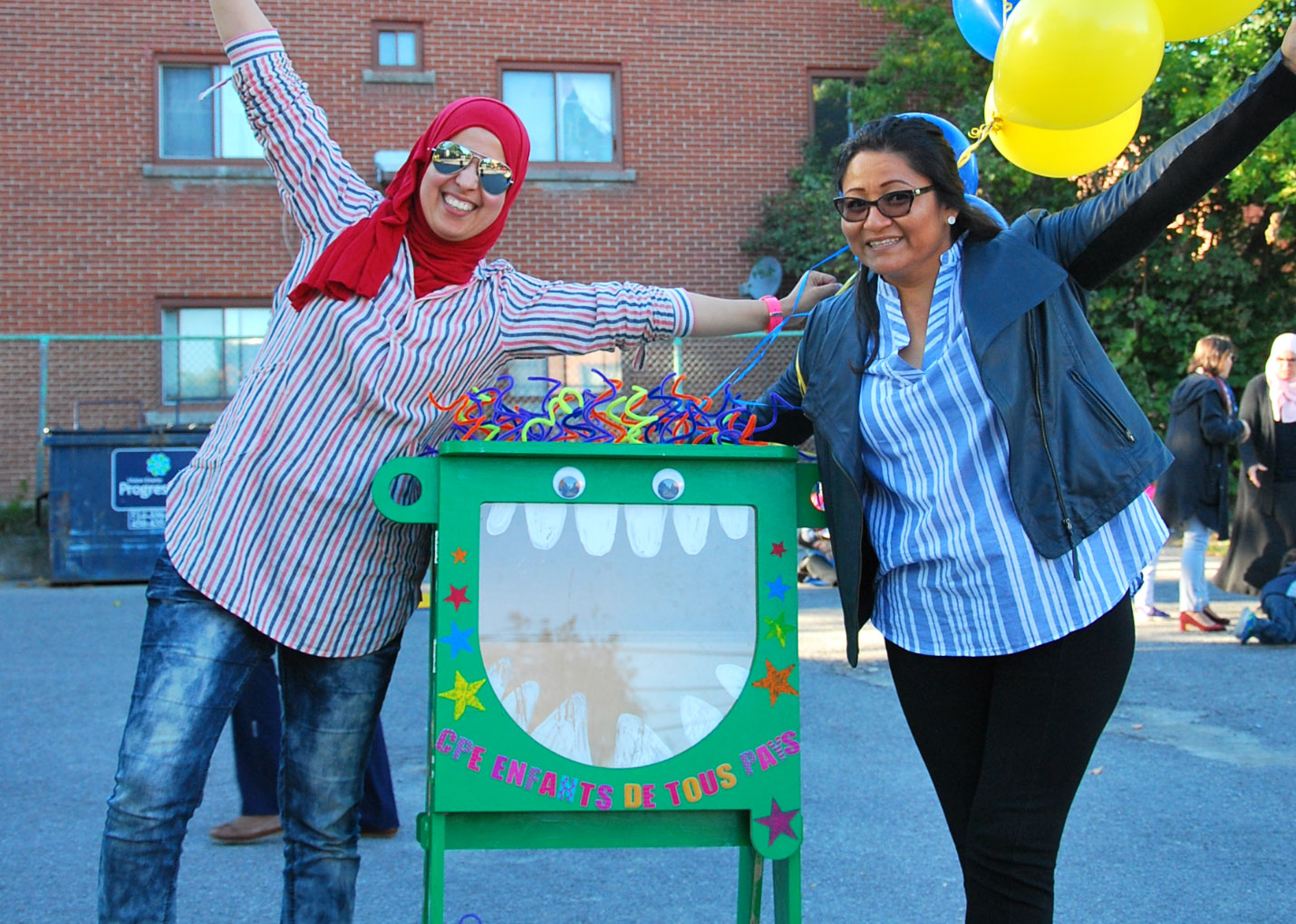 Deux femmes de Bordeaux-Cartierville se tiennent debout souriantes et bras ouverts avec des ballons autour d'un croque-livre (petite boite en bois ou tous le monde peut déposer et prendre des livres).